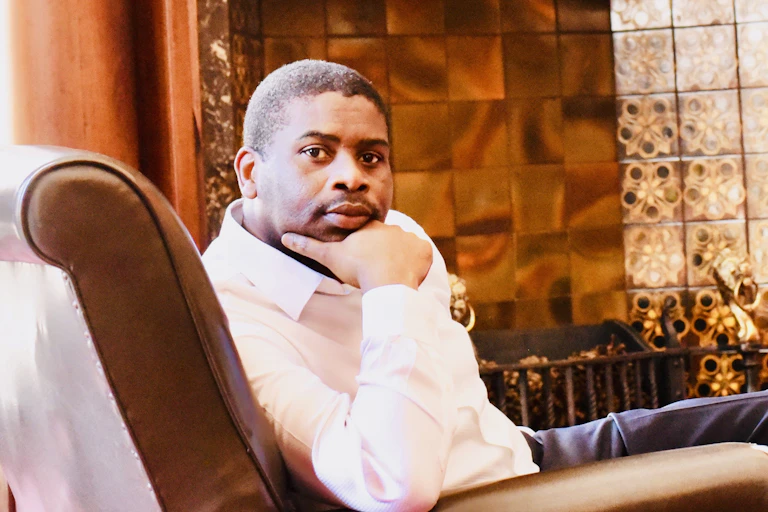 A man in a white shirt sits in a leather chair, his hand around his chin, looking directly to camera.