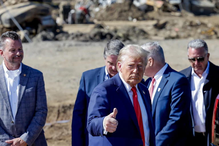 Donald Trump in a blue jacket does a thumbs up in an area with hurricane damage.