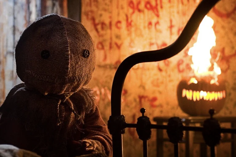 Small character with a burlap bag covering his head stands next to a jack 'o lantern that's on fire.