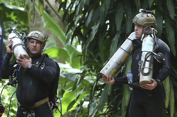 Two men in diving suits carry canisters of oxygen through a jungle