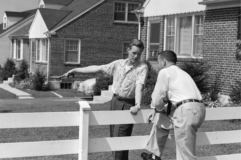 black and white photo of two men talking across a fence in a suburban neighborhood
