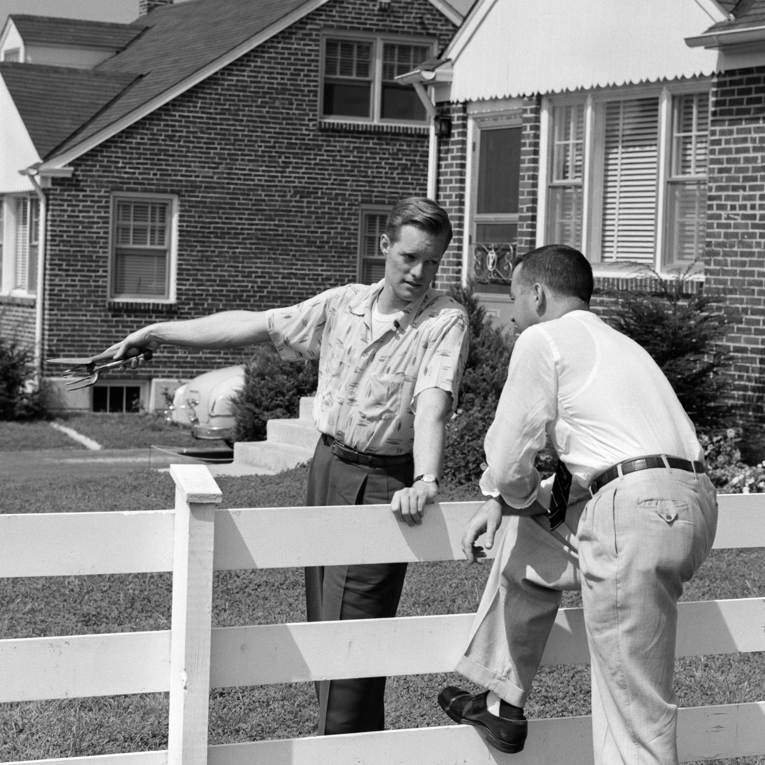 black and white photo of two men talking across a fence in a suburban neighborhood