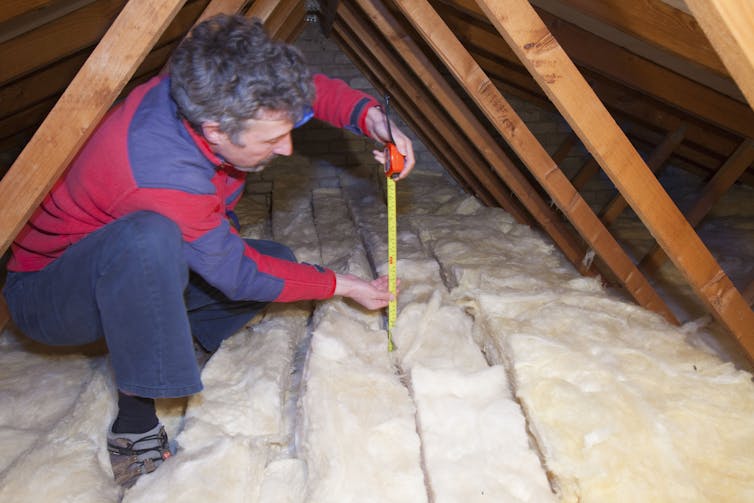 A man measures the depth of insulation in a house's small attic using a tape measure.