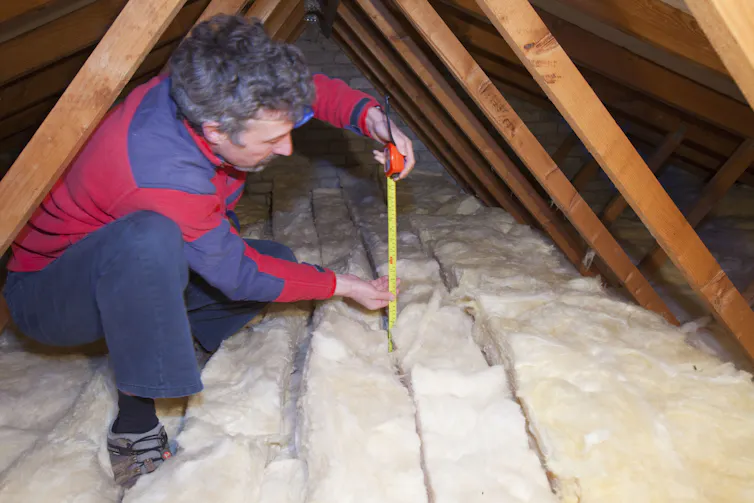 A man measures the depth of insulation in a house's small attic using a tape measure.