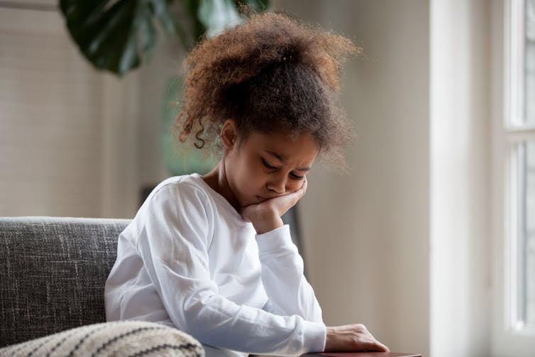 A young girl sits on a couch with her head in her hand.