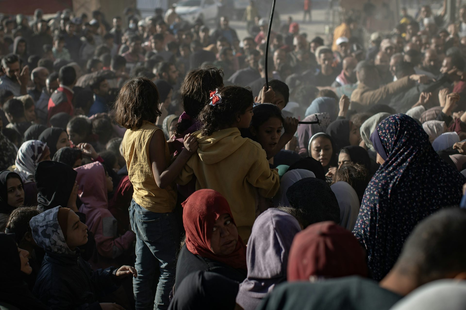 Three little girls stand up in a sea of people waiting for aid.