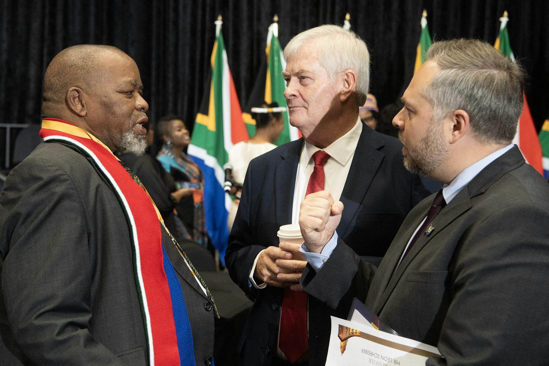 Three men wearing suits chat with in front of South African flags.