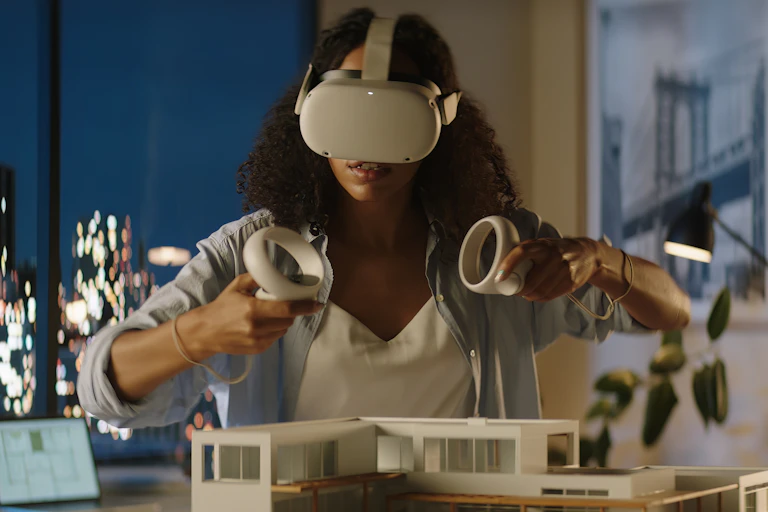 Une femme travaille avec un casque de réalité virtuelle et des manettes. Devant elle, une maquette d'architecte.