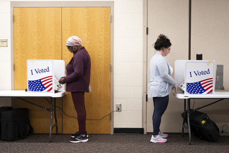 Two people stand facing away from each other at tables surrounded by small barricades to prevent people from peeking at their votes.