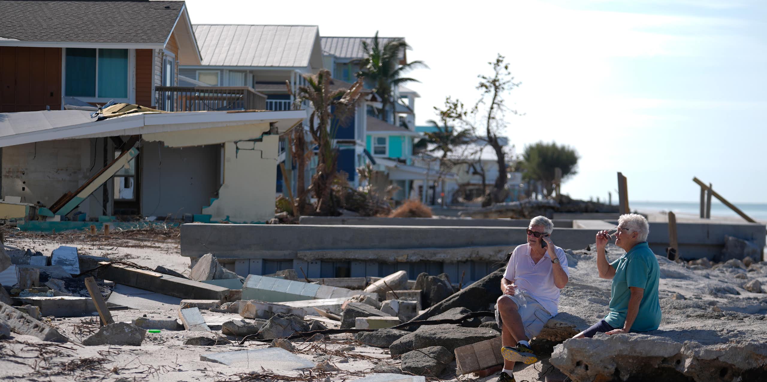 A man and woman sit on rubble on a beach near damaged homes