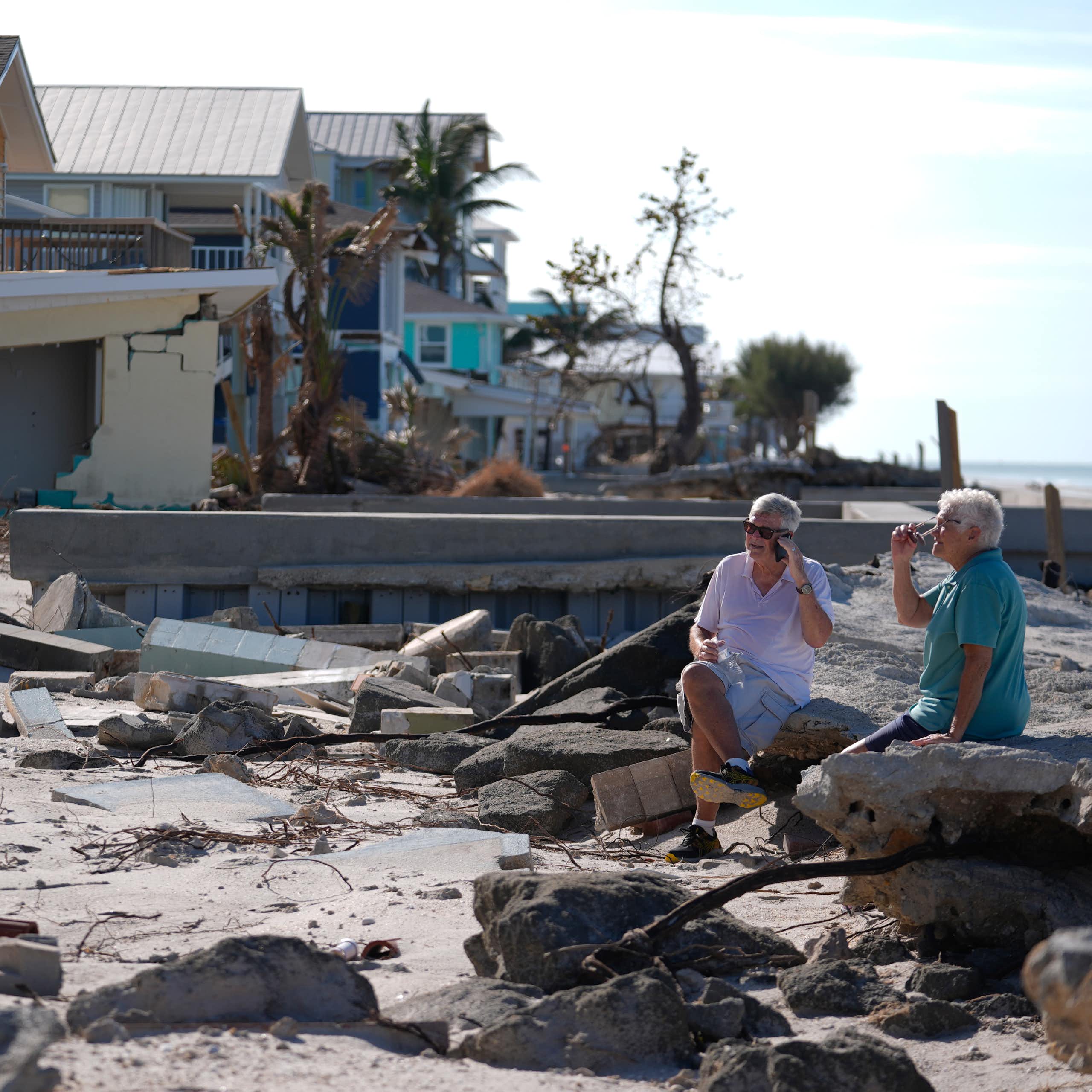 A man and woman sit on rubble on a beach near damaged homes