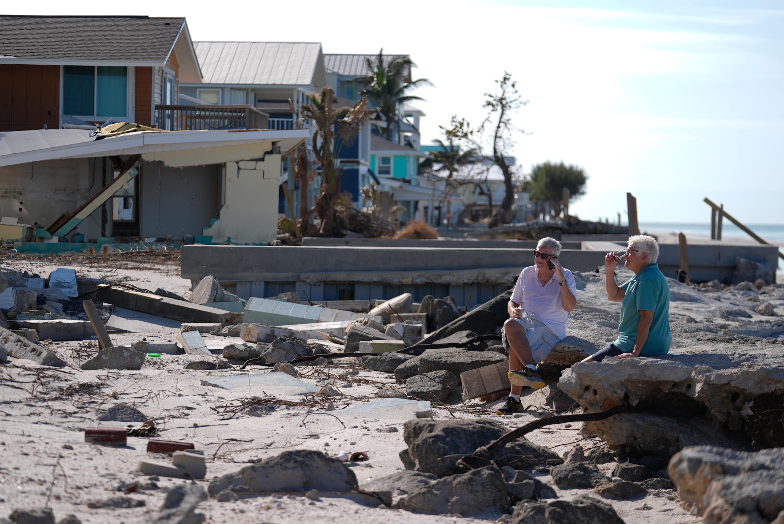 A man and woman sit on rubble on a beach near damaged homes
