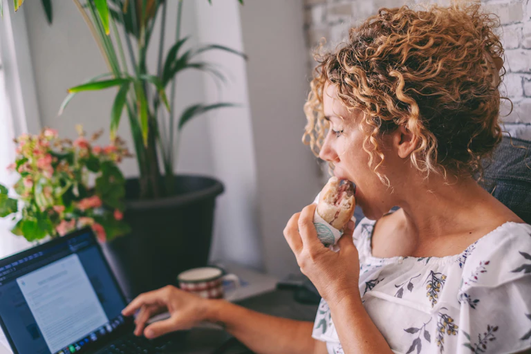 Mulher comendo um sanduíche enquanto trabalha no computador.
