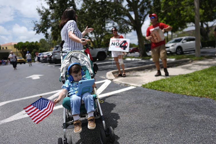 A woman with her back to the camera gestures toward two figures in the background with signs. In the foreground is a young child in a stroller listening to headphones and waving an American flag.
