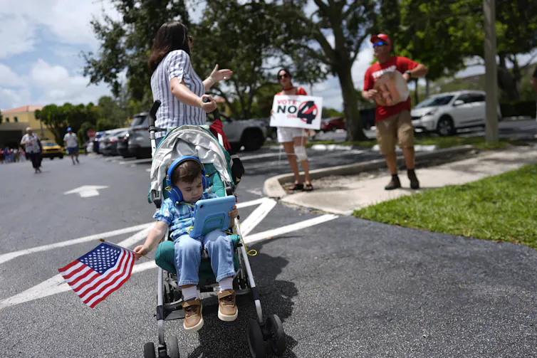 A woman with her back to the camera gestures toward two figures in the background with signs. In the foreground is a young child in a stroller listening to headphones and waving an American flag.