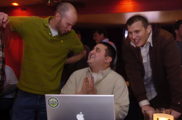 A man in a green golf shirt shares a happy look with a man who is seated in front of a laptop.