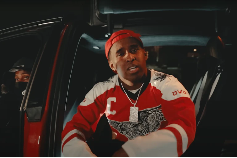 A Black man wearing a red sport jersey and baseball cap sits in the back seat of a car looking into the camera.