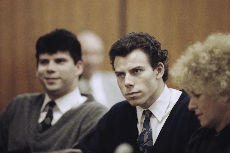 Two men in shirt and tie sit in a court room with a woman in a dark jacket