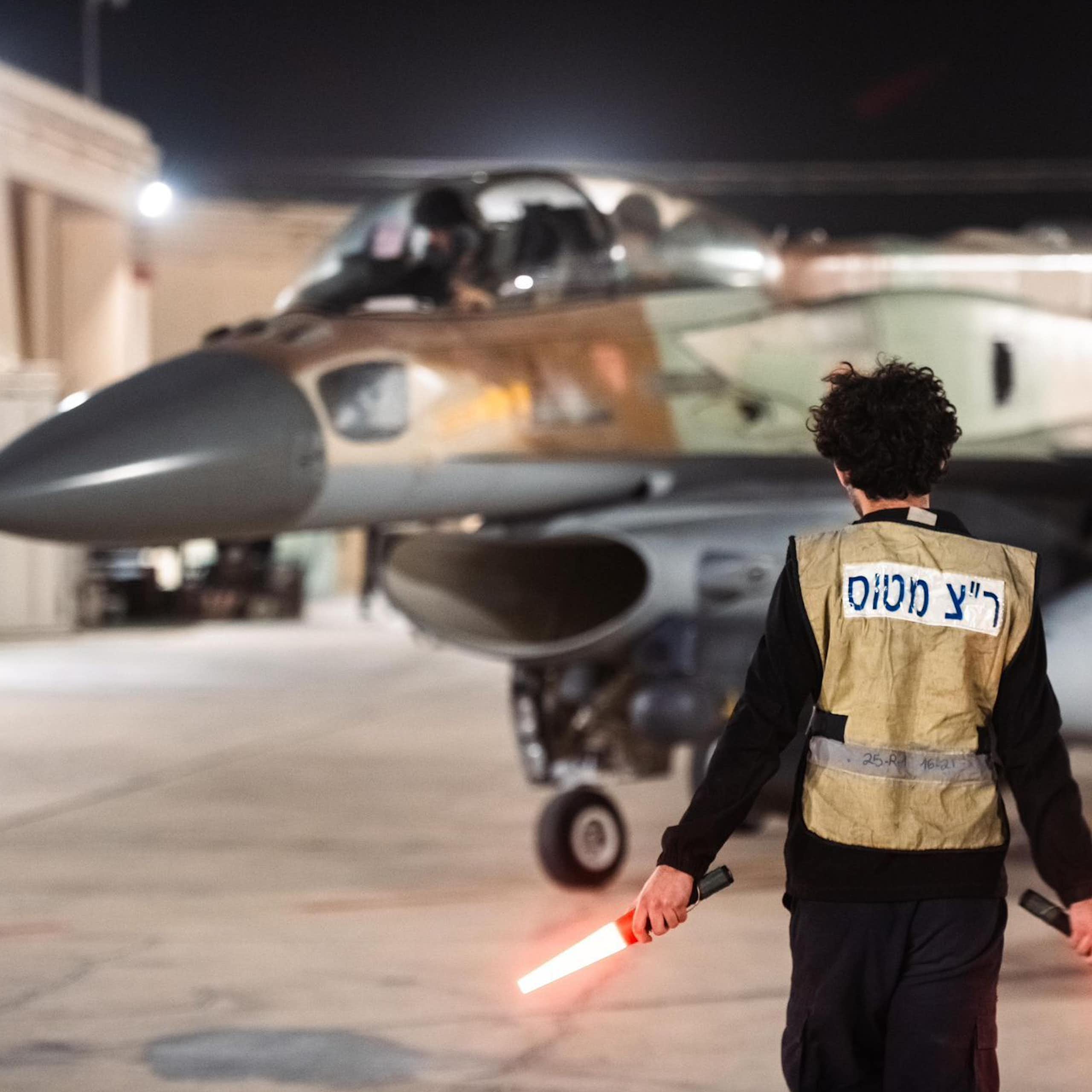 A man with hand-held lights stands in front of a military jet.