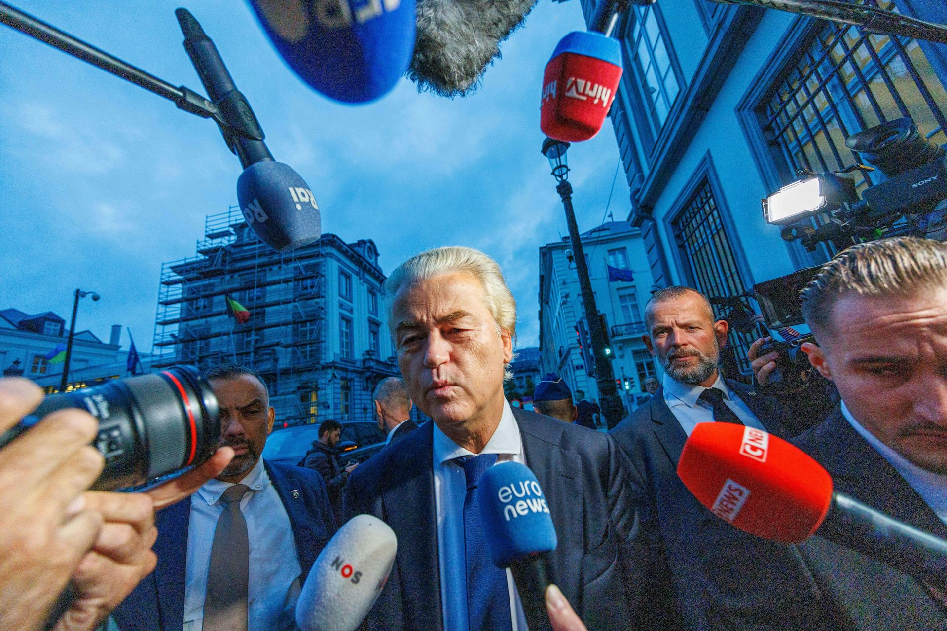 Geert Wilders walking down a street surrounded by press microphones.