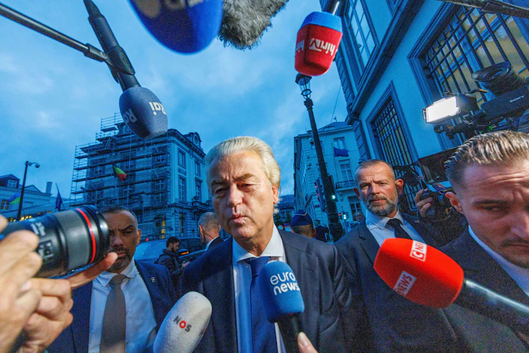Geert Wilders walking down a street surrounded by press microphones.