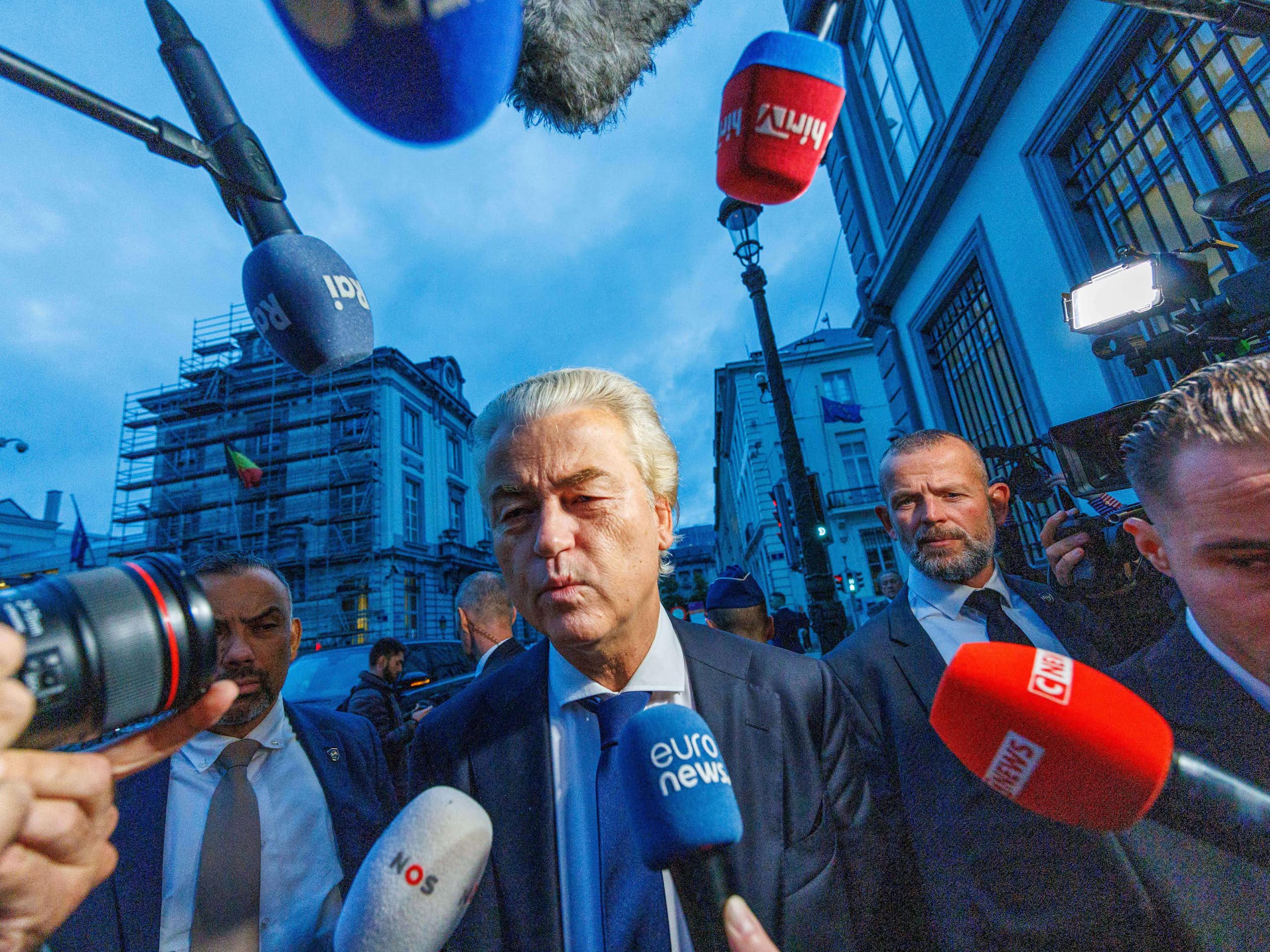 Geert Wilders walking down a street surrounded by press microphones.