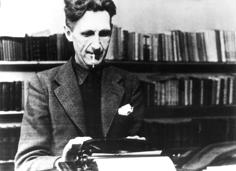 A dark haired man wearing a sport jacket and smoking a cigarette while working at a typewriter.