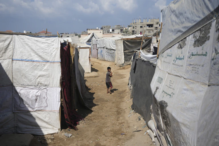 A small boy carries a small loaf of bread while walking between white makeshift tents.