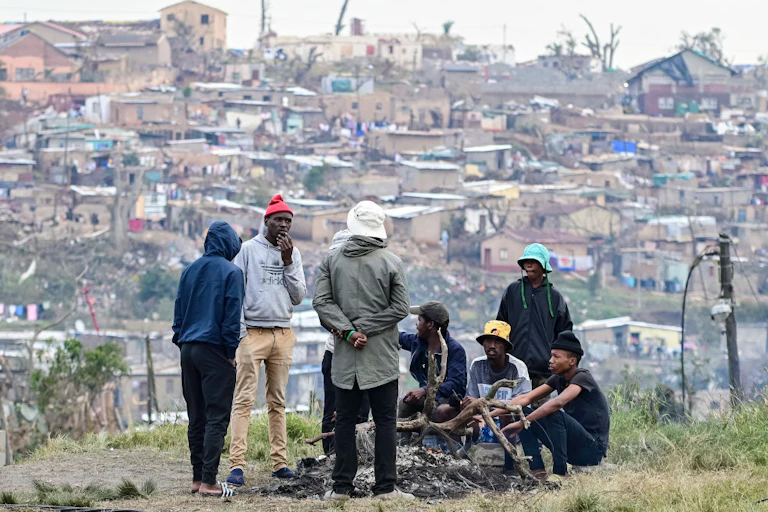 Young men around a fire on open ground. A dense settlement in the background.