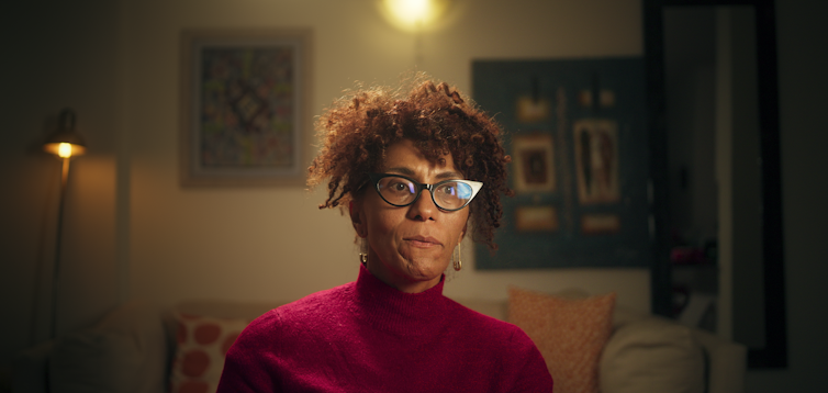 A woman in a burgundy polo neck shirt with horn-rimmed glasses talks to camera.
