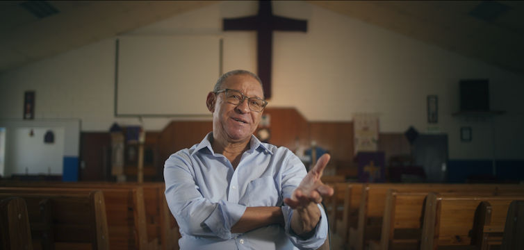 A balding man with glasses gestures with one hand as he talks to camera, behind him a large cross on a wall.