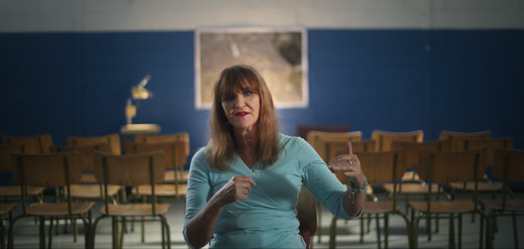 An older woman in a blue dress gestures with both hands as she talks to camera, behind her a classroom with an overhead projector.