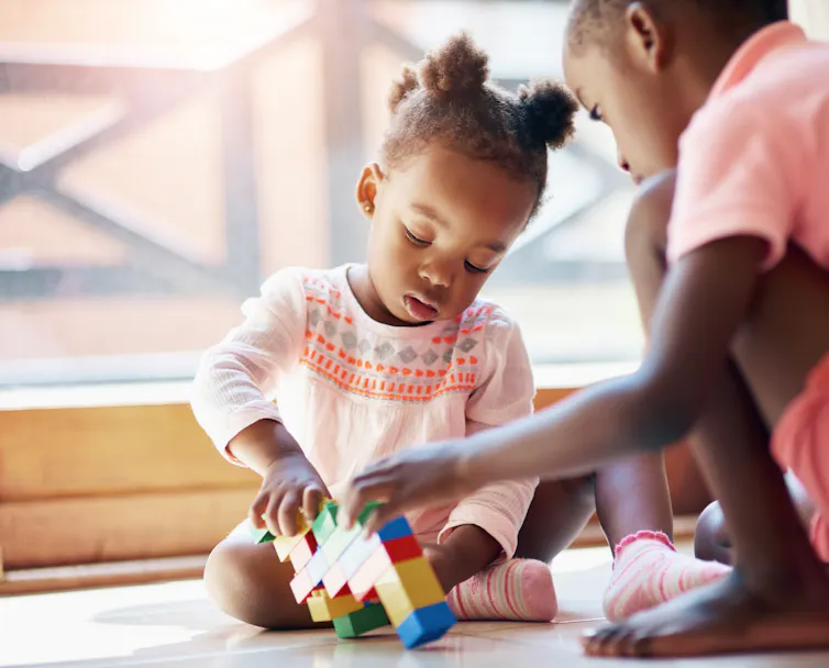 A little girl plays with an older sibling or friend. They are fitting building blocks together.
