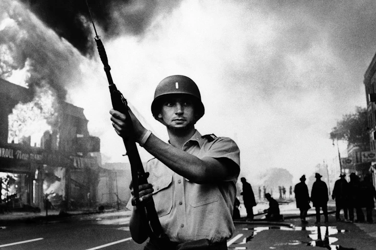 A soldier holding a rifle watches over firemen battling a fire.