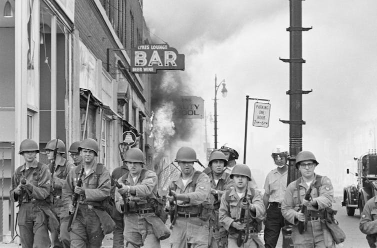 Soldiers with rifles guard a burning building.