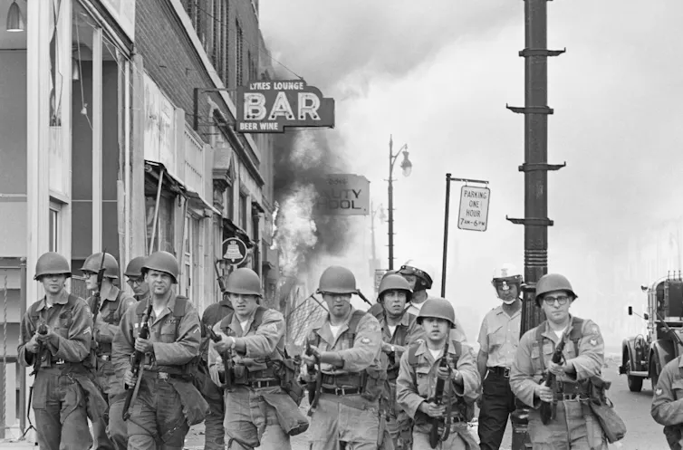 Soldiers with rifles guard a burning building.