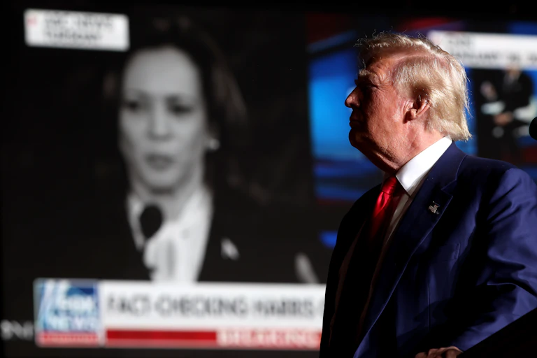 A man in a blue suit stands looking at a big screen with a woman's face on it.
