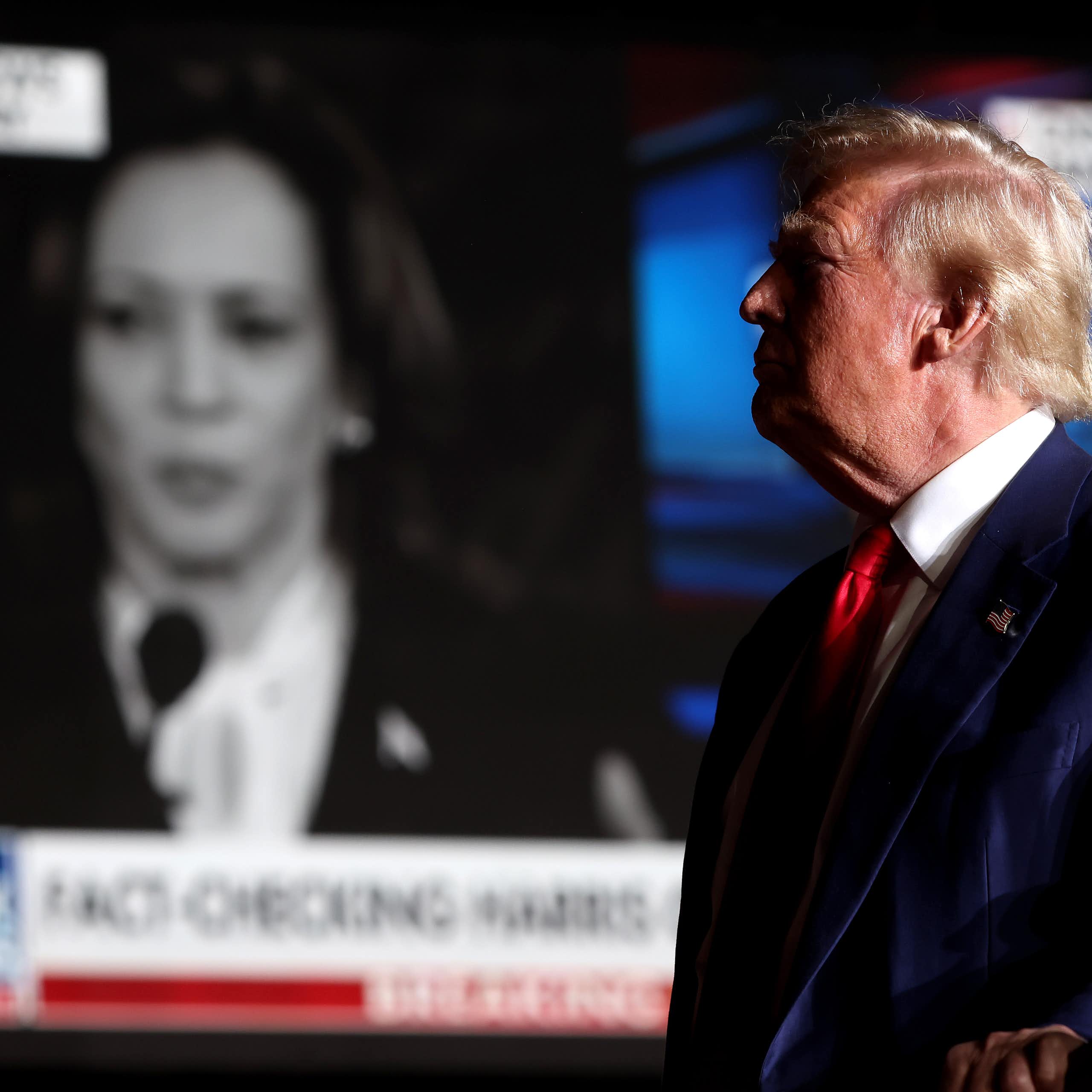 A man in a blue suit stands looking at a big screen with a woman's face on it.