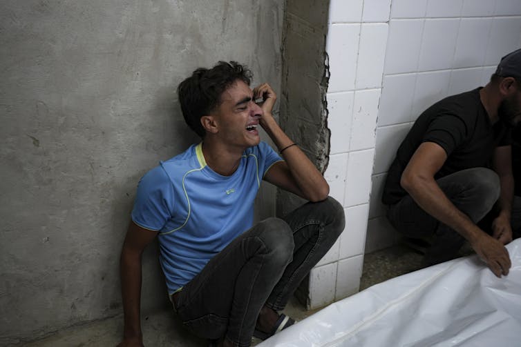 A boy in a blue t-shirt sits against a wall at a hospital sobbing.