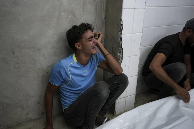A boy in a blue t-shirt sits against a wall at a hospital sobbing.