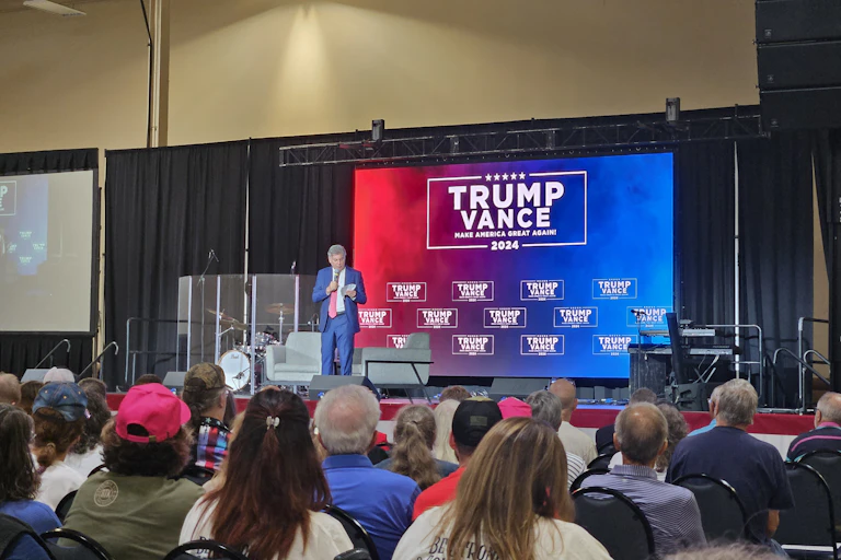 A man dressed in a blue suit speaks into a microphone to an audience seated in front. On the splash screen behind him are names of Trump and Vance.