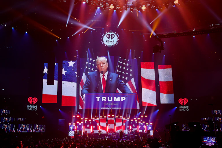 A white man with a blue suit stands at podium in a large sark room, with screens on the side and in front showing sliced bits of the American flag. The word 'Trump' is on the podium.