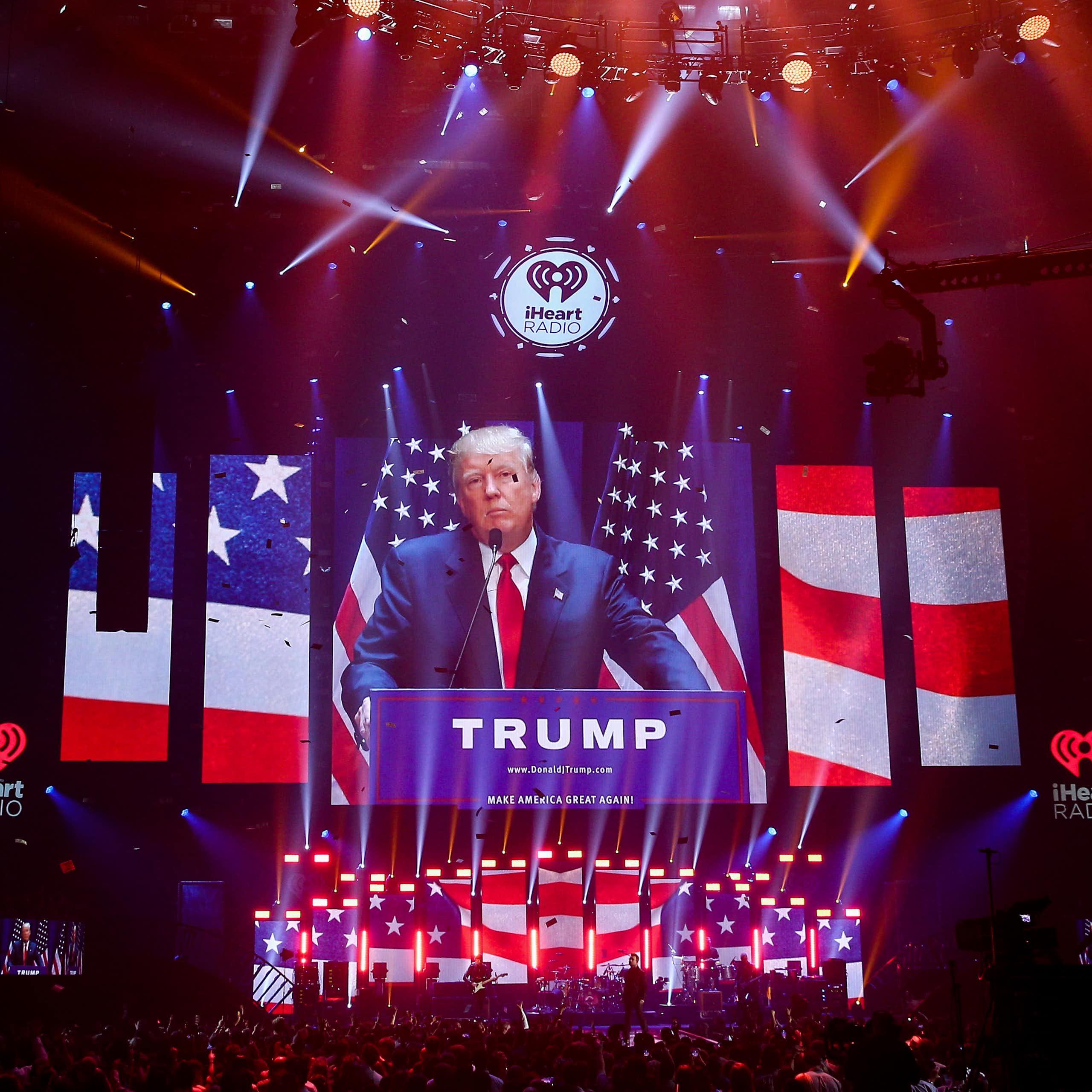 A white man with a blue suit stands at podium in a large sark room, with screens on the side and in front showing sliced bits of the American flag. The word 'Trump' is on the podium.