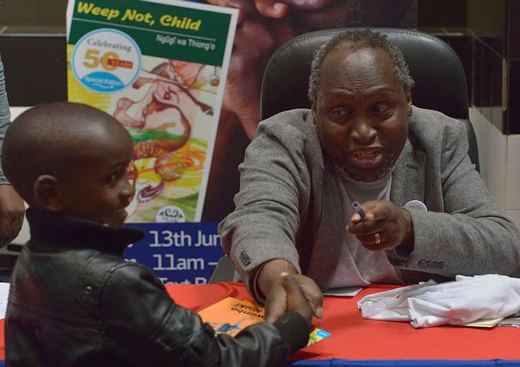 An older man sits at a table where he is signing books, a boy stands in fr ont of the table and shakes his hand.