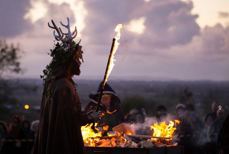 Bearded man wearing antlers holds a torch in front of a bonfire as other people look on.
