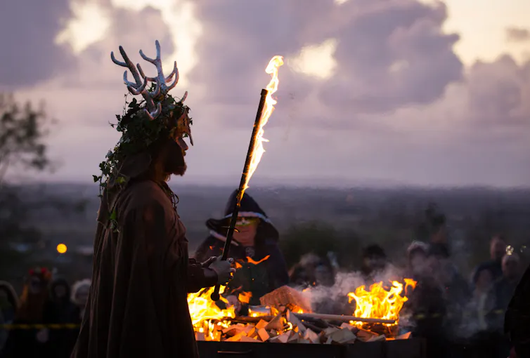 Bearded man wearing antlers holds a torch in front of a bonfire as other people look on.