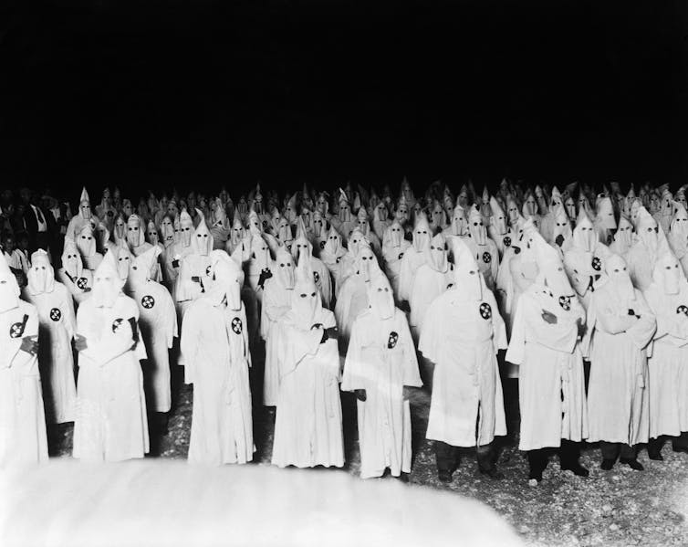 Dozens of people wearing white robes and hoods are standing together on a field.