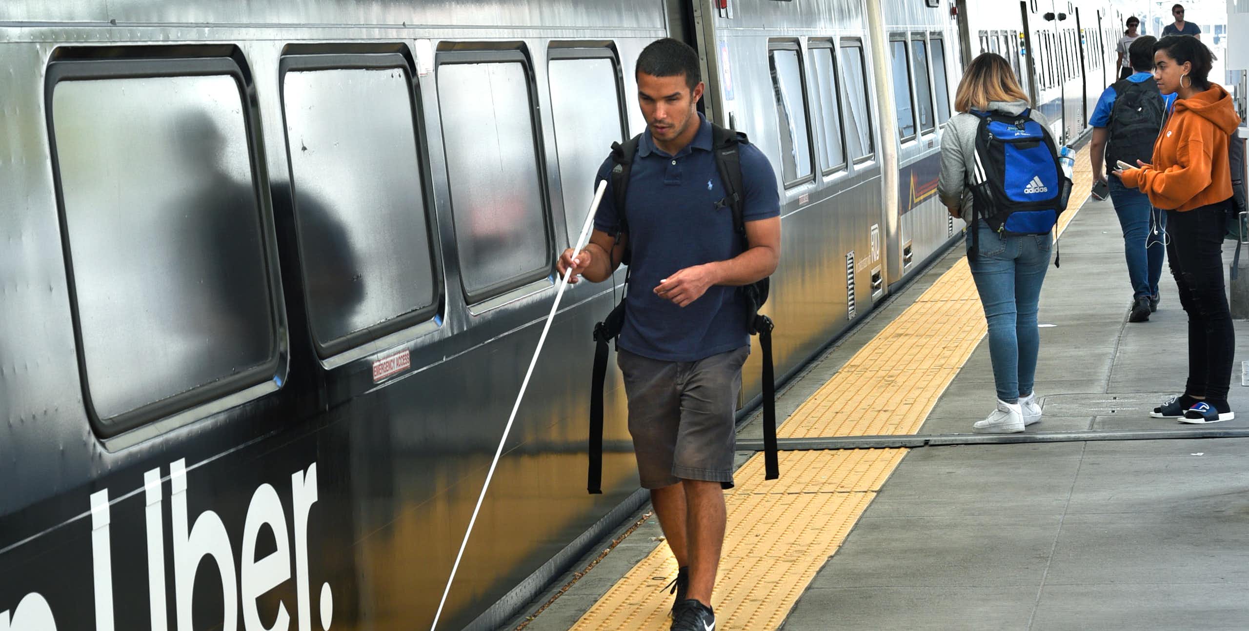 A young blind man walks alongside a train, using a thin white cane.