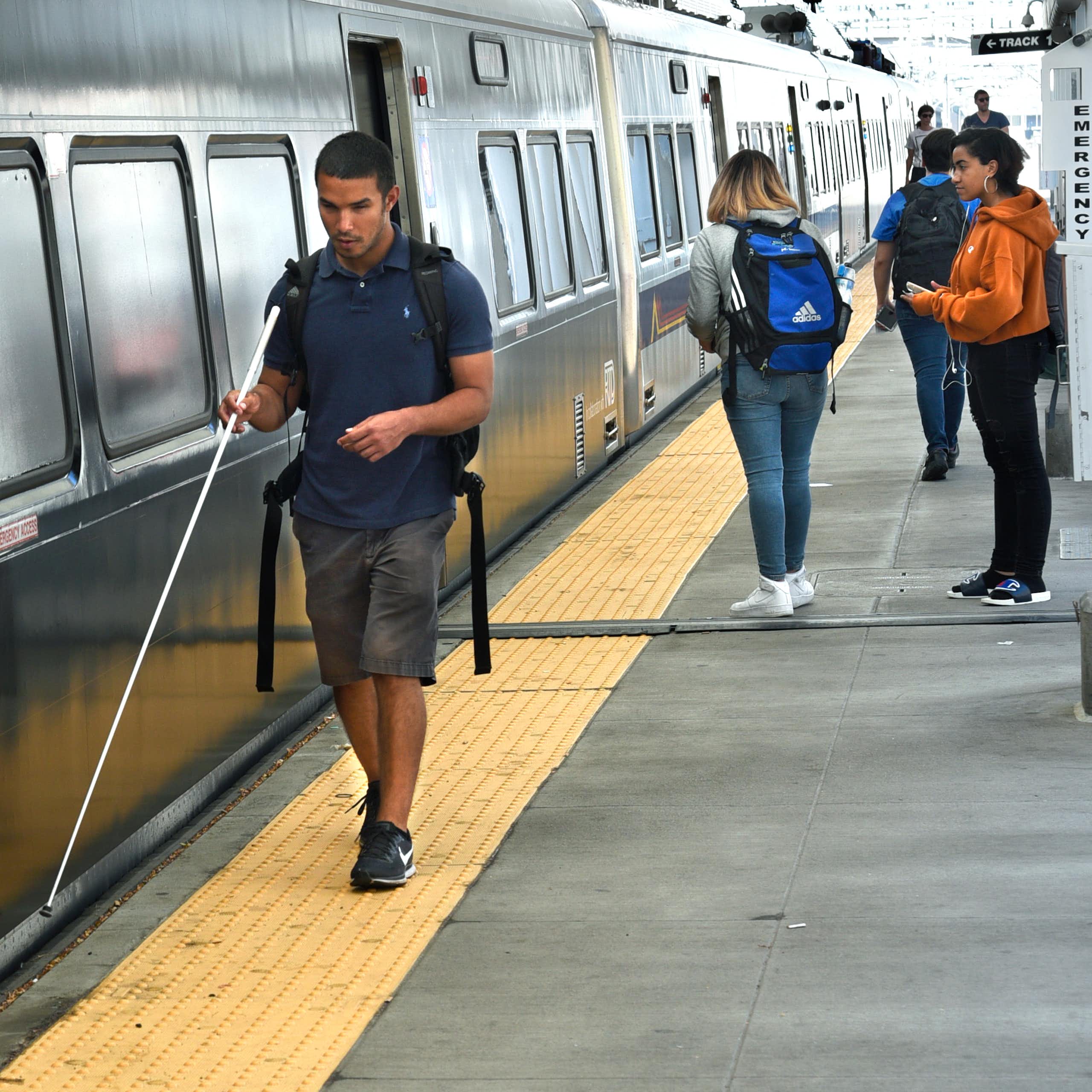 A young blind man walks alongside a train, using a thin white cane.