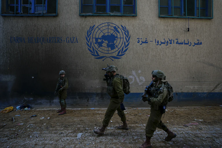 Soldiers in fatigues and helmets walk past a building with a UN logo on it.
